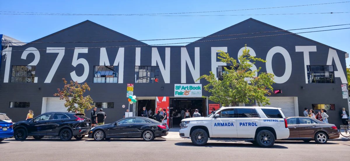 A white security SUV labeled “ARMADA PATROL” parked outside the SF Art Book Fair entrance at 1275 Minnesota Street, while attendees gather near the doors.